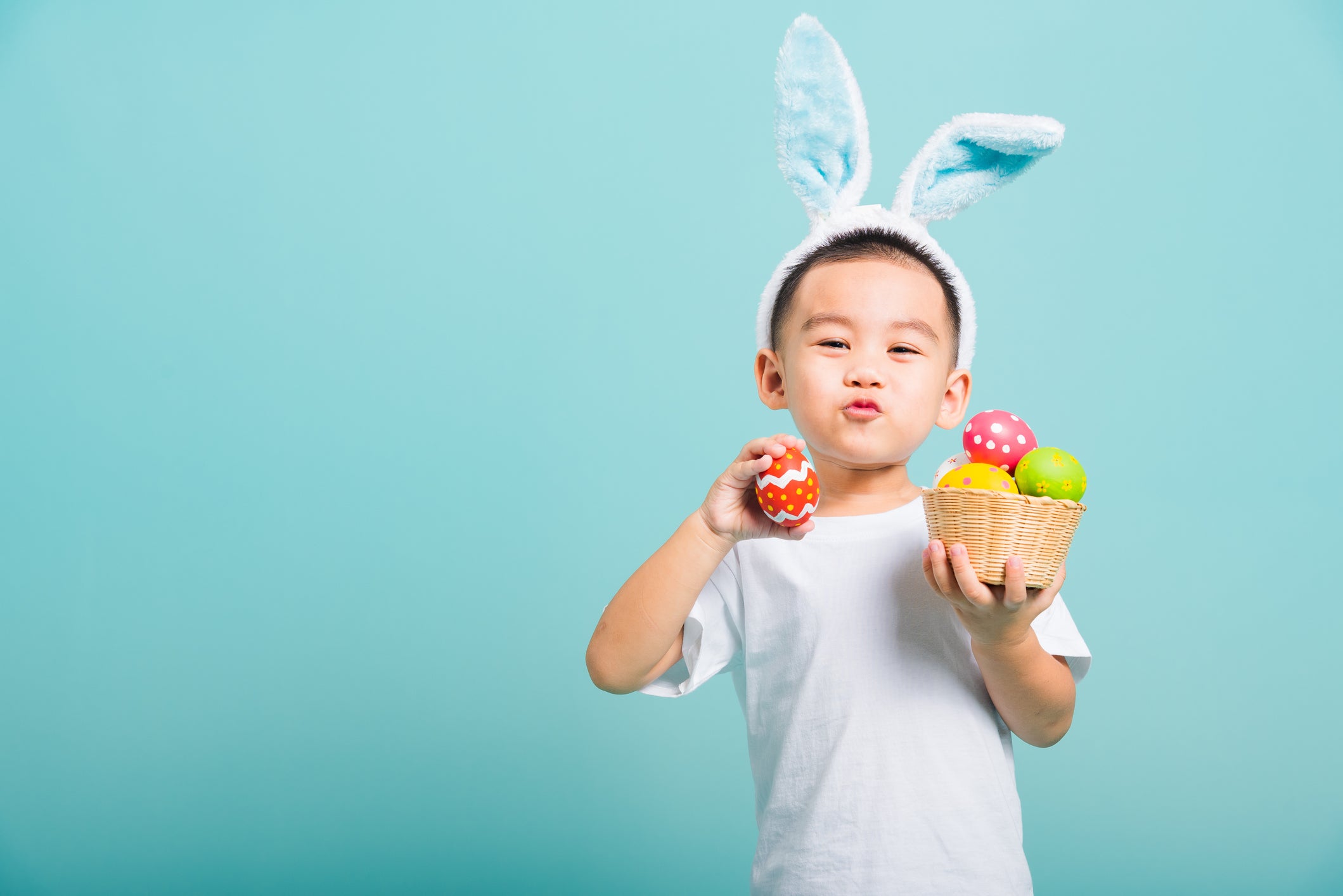 Asian cute little child boy smile beaming wearing bunny ears and a white T-shirt, standing to hold a basket with full Easter eggs. And other hand holds an easter egg